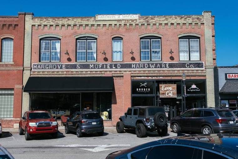 A three-story brick building with a sign reading 'Hargrove &amp; Mofield Hardware Co.' on the upper facade, with three large windows on the second floor and storefronts on the ground level. Several parked cars, including a red SUV and a black Jeep, are in front of the building on a parking lot under a clear blue sky.