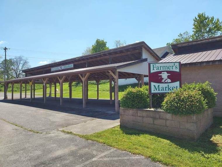 Exterior of a rural farmer's market with a covered parking area, green shrubbery, and a sign reading "Farmer's Market" featuring a farmer silhouette