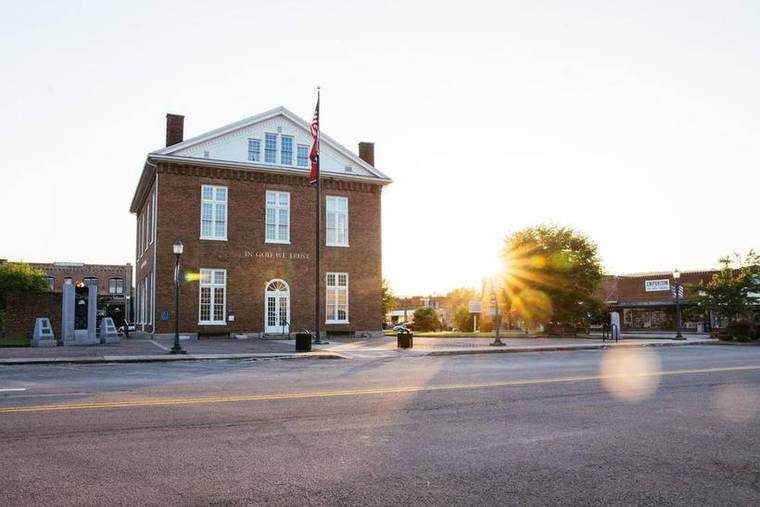 A red brick building with white trim and multiple windows, a flagpole with the American flag, and the words 'In God We Trust' on the brick wall. The sun is setting behind a tree, casting a warm glow and lens flare. Streets and sidewalk are in the foreground.