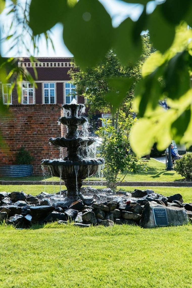 A decorative fountain surrounded by rocks in a park with green grass, trees, and a brick building in the background.
