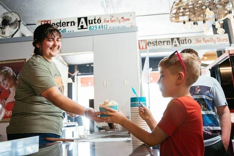 A young woman handing a cup of ice cream to a boy at an ice cream shop. The shop has signs for Western Auto and a stack of cups on the counter.