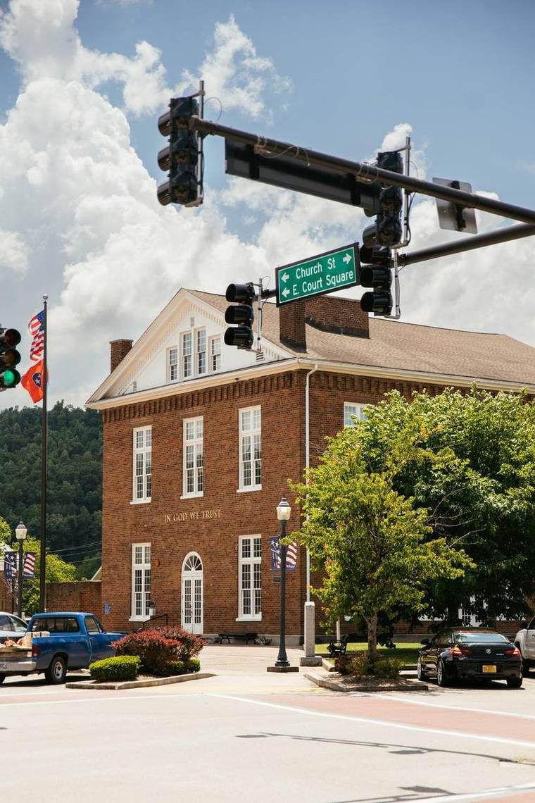 A brick building with white trim and the words 'In God We Trust' inscribed on it. There are American flags and other banners around. Traffic lights and street signs indicate the intersection of Church Street and East Court Square. Several cars and trees are in the foreground, set against a sky with white clouds.