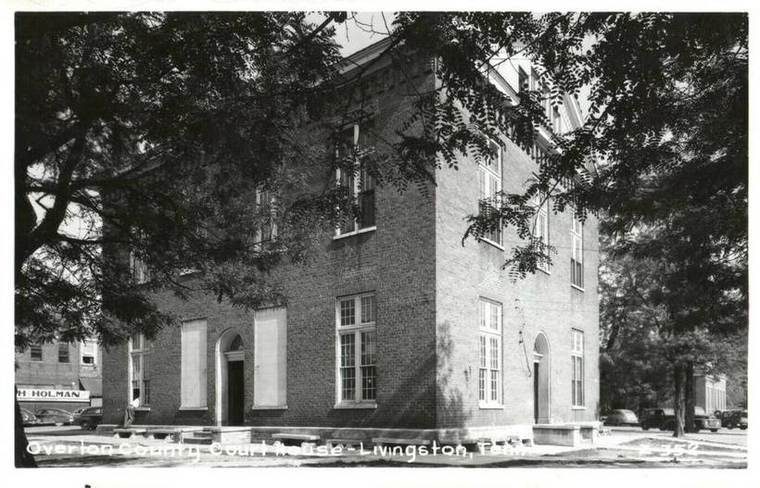 Black and white photo of a brick courthouse building with tall windows and an arched doorway, surrounded by trees and parked cars in the background.