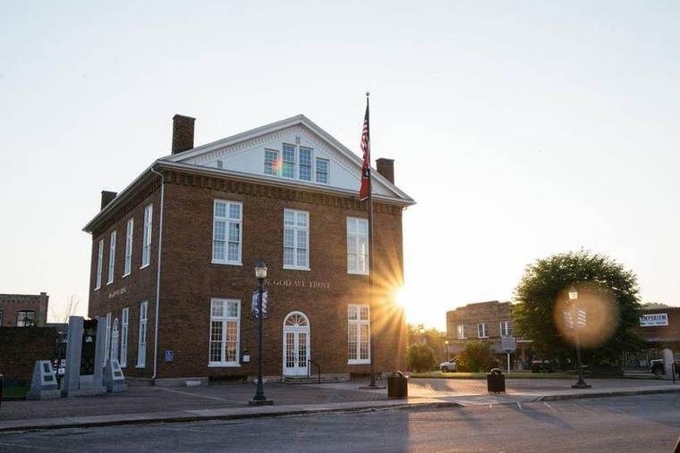 A brick historic courthouse with white windows and a decorative white gable at the top, backlit by the setting sun. An American flag is flying in front of the building, and there are street lamps, benches, and a few trees in the surrounding area.