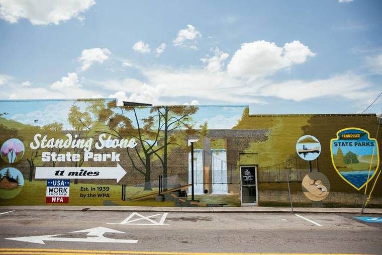Colorful mural on a brick wall featuring trees, a lake, and outdoor scenes. A sign indicates the location is Standing Stone State Park, Tennessee, 11 miles away, established in 1939, with references to the WPA work program.