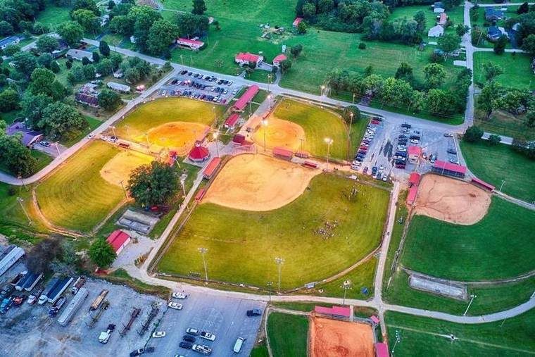 Aerial view of a baseball complex with multiple baseball fields illuminated at dusk, surrounded by parking lots, residential houses, and trees.