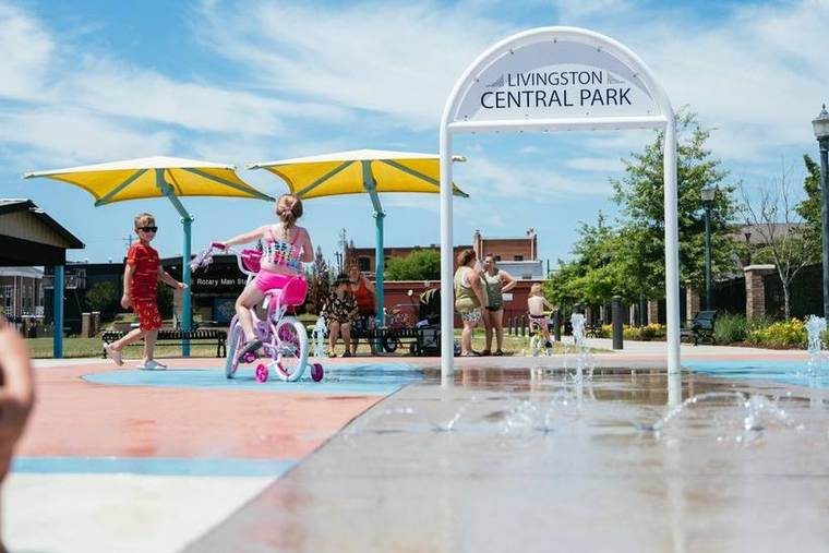 Children playing in a splash pad at Livingston Central Park with yellow umbrellas and people sitting on benches.