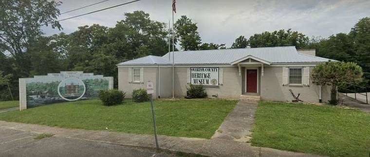 A small, light-colored building with a metal roof, labeled as Overton County Heritage Museum. There is a sign on the front of the building, a flagpole with an American flag, a small path leading to the entrance, and a painted mural on the left side depicting a historic scene. Green grass and trees are in the background.