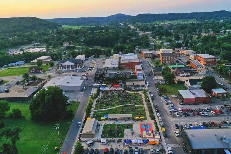 Aerial view of a small town during a music concert with a stage at the center, surrounded by a crowd, food stalls, and a playground with basketball courts, set against a backdrop of green hills and trees at sunset.