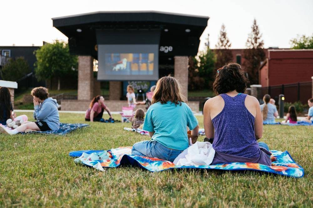 People sitting on blankets watching an outdoor movie on a large screen in a park during evening with trees and buildings in the background.